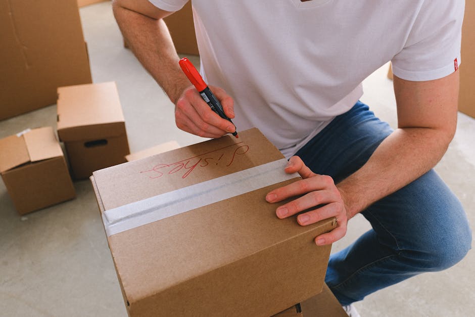 A person wearing a white t-shirt and blue jeans is kneeling inside a room filled with cardboard boxes used for house removals. They are holding a red marker in their right hand and writing on a medium