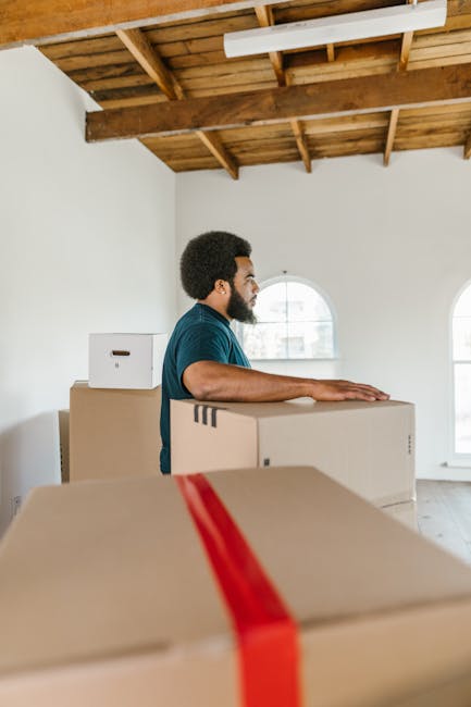A person wearing a white t-shirt and blue jeans is kneeling inside a room filled with cardboard boxes used for house removals. They are holding a red marker in their right hand and writing on a medium