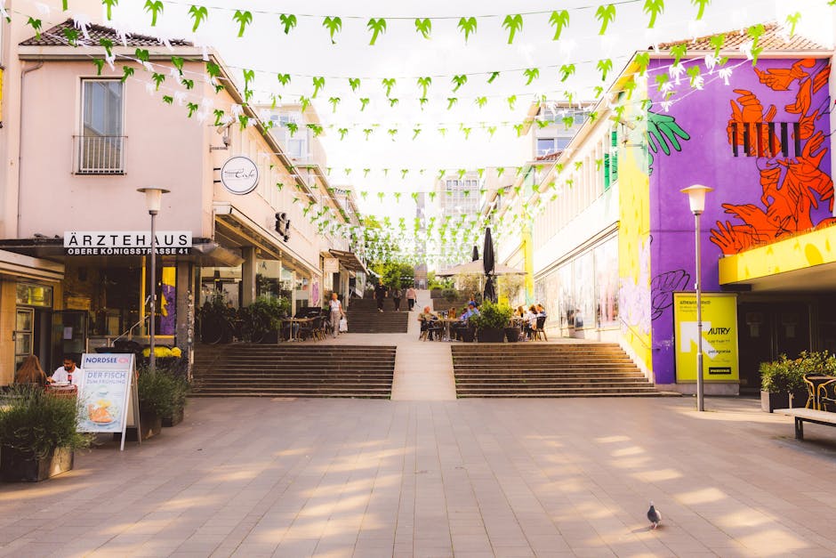 A wide view of a pedestrian shopping area with multiple shops and cafes on either side, featuring potted plants and outdoor seating. The area is connected by stone steps leading up to an open space decorated with overhead green and white hanging banners. Several people are visible walking, sitting, and socialising around tables with umbrellas, while a pigeon walks across the paved ground in the foreground. Bright sunlight illuminates the scene, with inbuilt lighting fixtures on lamp posts. The surrounding buildings include shopfronts with signage, with some murals painted on the walls, and the general environment suggests a lively, pedestrian-friendly urban centre suitable for home relocation and furniture transport logistics. Movements of individuals carrying items or preparing for moving activities may be part of the scene, with potential involvement from professional movers like Movers Bromley for house removals and packing services.