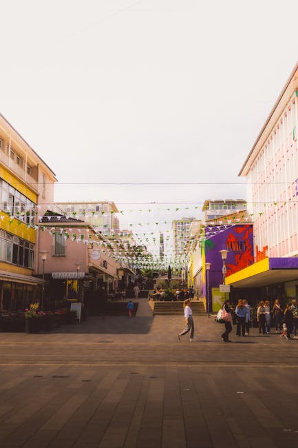 Wide-angle view of an outdoor shopping and entertainment area with colourful buildings on either side, featuring yellow, pink, and purple facades. String lights are hung overhead across the open space, which has a paved walkway where several pedestrians are walking, some carrying bags. The area appears to be part of a home relocation or furniture transport scene, with commercial storefronts and outdoor seating visible. Open staircases lead up to the buildings, and planters with greenery are placed along the pathways. The lighting is natural, with an overcast sky providing diffuse daylight, and the scene is associated with professional removals and moving services offered by Movers Bromley, supporting packing and logistics activities associated with house removals in Bromley.