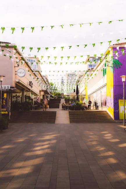 A wide view of a pedestrian shopping area with multiple shops and cafes on either side, featuring potted plants and outdoor seating. The area is connected by stone steps leading up to an open space decorated with overhead green and white hanging banners. Several people are visible walking, sitting, and socialising around tables with umbrellas, while a pigeon walks across the paved ground in the foreground. Bright sunlight illuminates the scene, with inbuilt lighting fixtures on lamp posts. The surrounding buildings include shopfronts with signage, with some murals painted on the walls, and the general environment suggests a lively, pedestrian-friendly urban centre suitable for home relocation and furniture transport logistics. Movements of individuals carrying items or preparing for moving activities may be part of the scene, with potential involvement from professional movers like Movers Bromley for house removals and packing services.