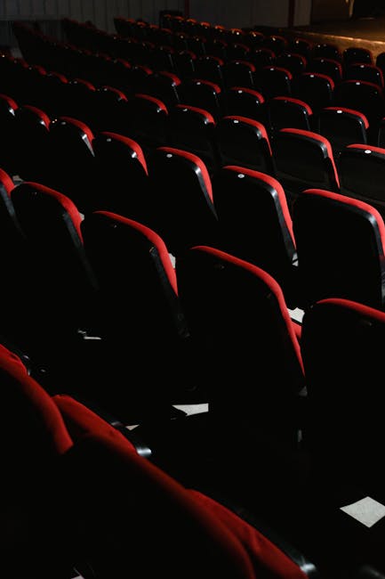 Interior view of the Churchill Theatre in Bromley, showing a stage with closed red velvet curtains illuminated by stage lighting, surrounded by rows of empty black theatre seats with red accents. The 