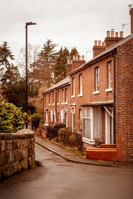 A narrow residential street with a slight incline, featuring a stone wall on the left side and a pavement on the right side. The street is lined with red brick and yellow brick houses with chimneys, d
